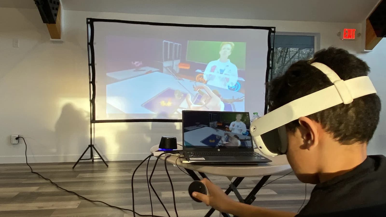 A high-school-age student at a table in a VR headset, seen from behind, his lesson view mirrored on the screen behind him: a rendered classroom with an avatar in it.