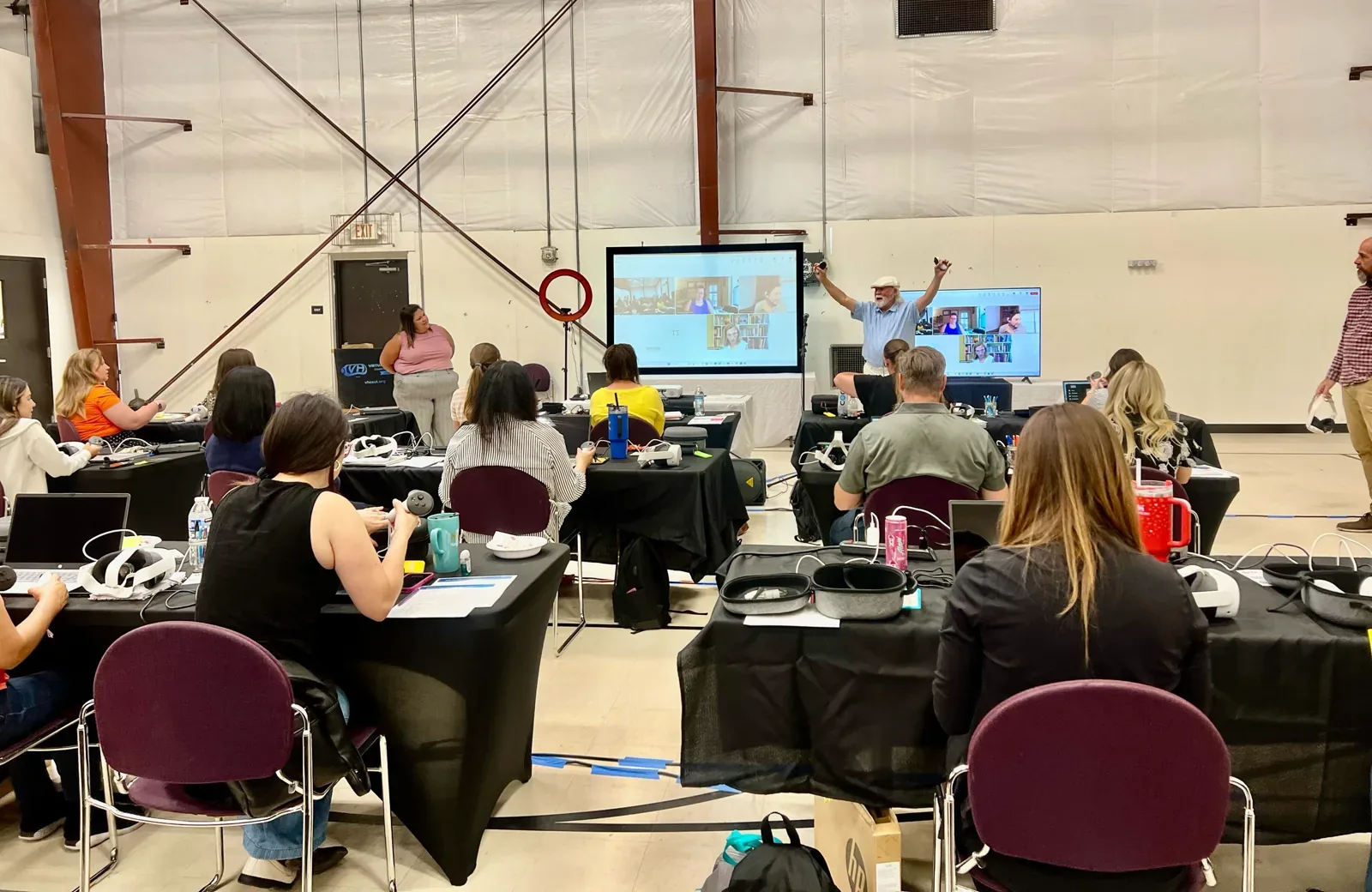 A training hall of teachers at long tables with laptops and Quest headsets, facing a presenter and a screen carrying a video call